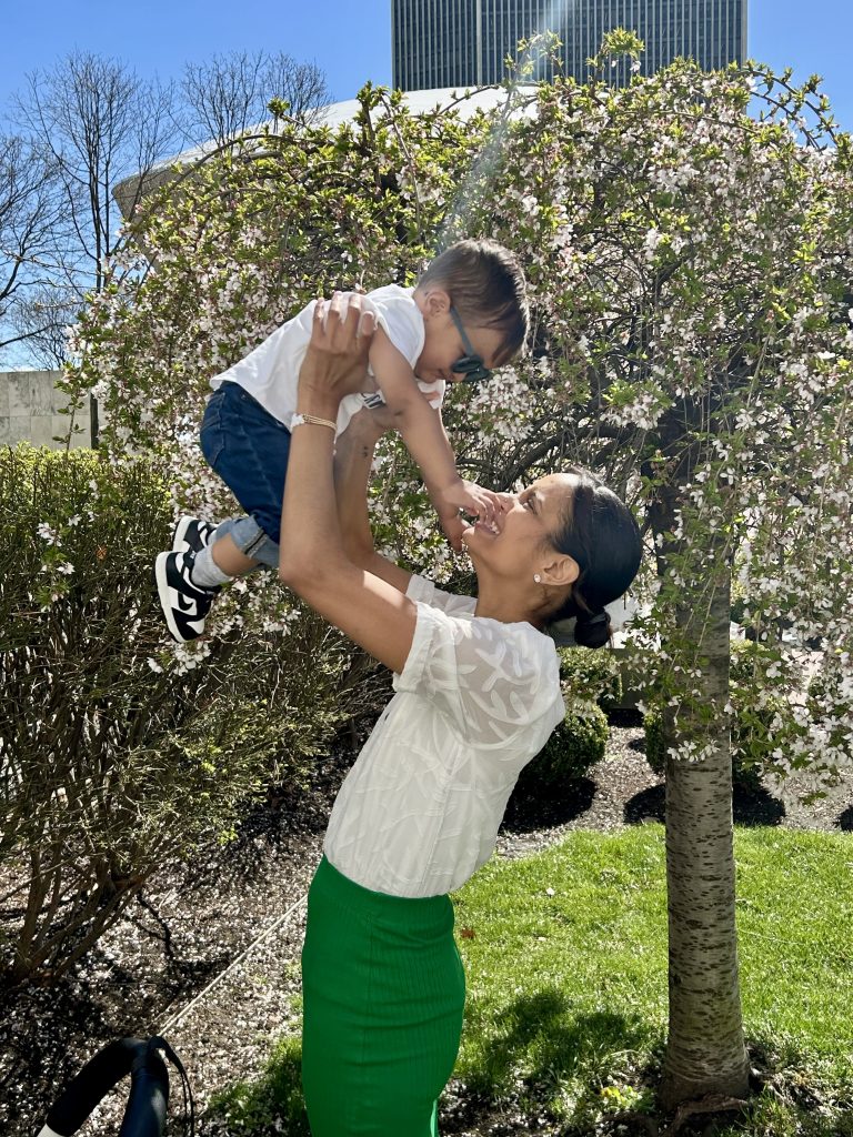 A woman in a white shirt and green skirt holds a baby up above her head. The baby has glasses, a white shirt, jeans and sneakers on.